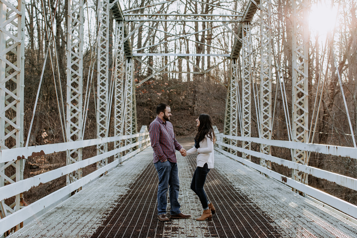 Happy couple, holding hands, standing in the center of a white, rustic looking bridge with industrial feel and tones. 
