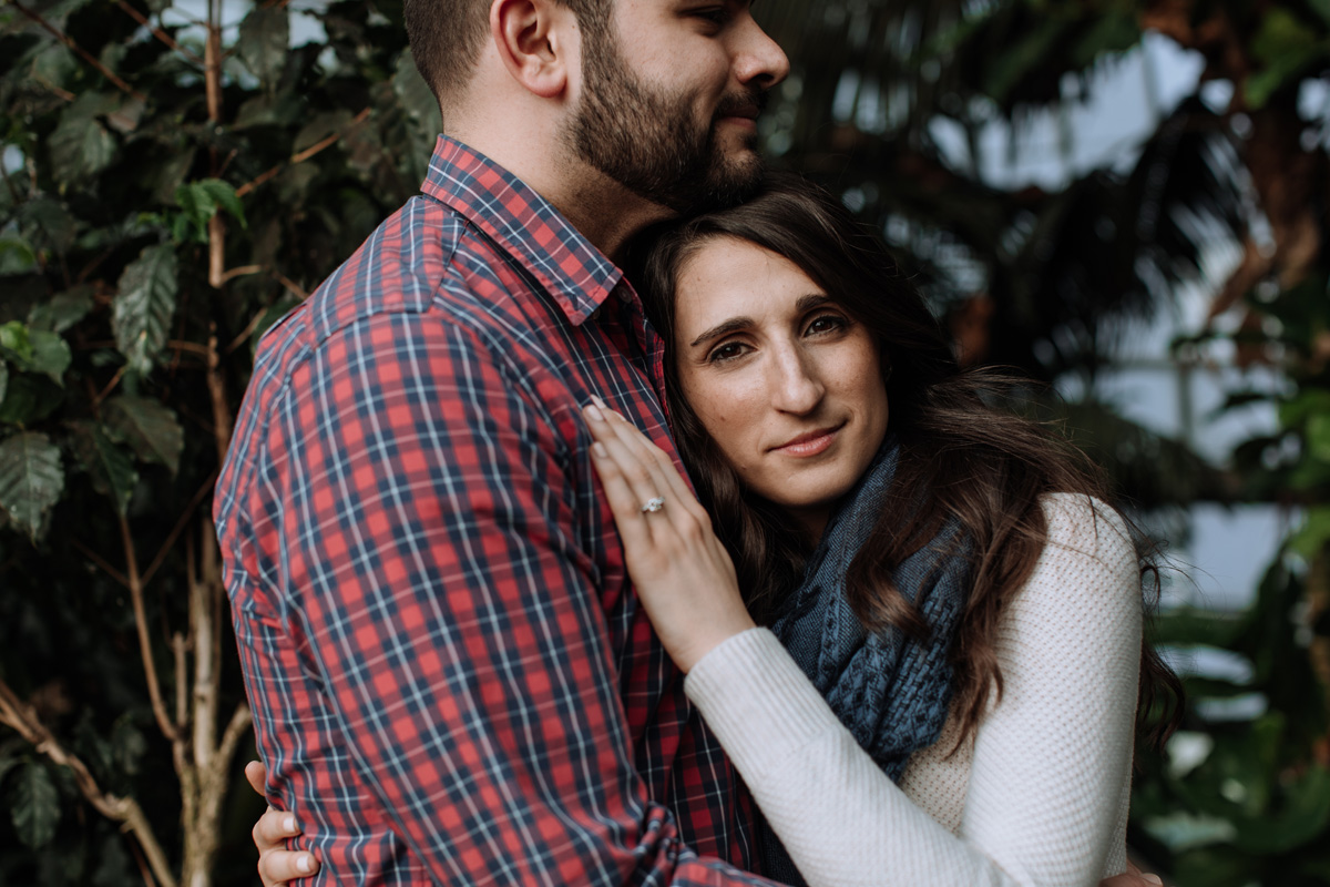 Young couple embracing each other with man looking to the right, and woman looking forward towards camera with her hand resting on man's chest. Man has a scruffy beard and is wearing a red, white, and dark blue plaid button down shirt. Woman has engagement ring on finger of left hand while her hand rests on man's chest, while wearing a white longsleeve shirt and bluish green scarf.