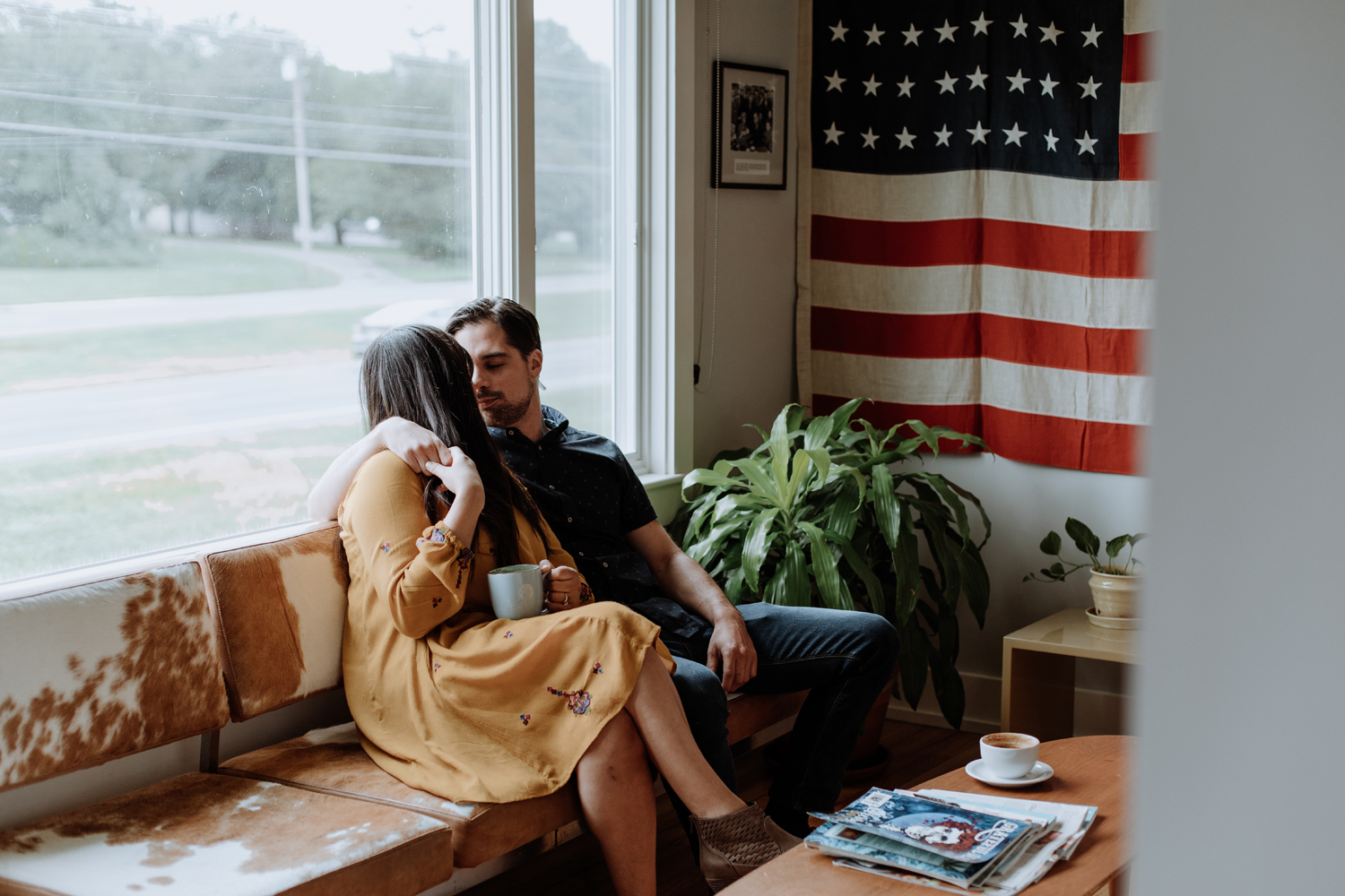 Casually dressed couple embracing each other, staring intently into one another's eyes, while sitting on a vintage brown and white-colored couch overlooked by a red, white, and blue United States of America flag hanging on the wall to the far right side of the photo.