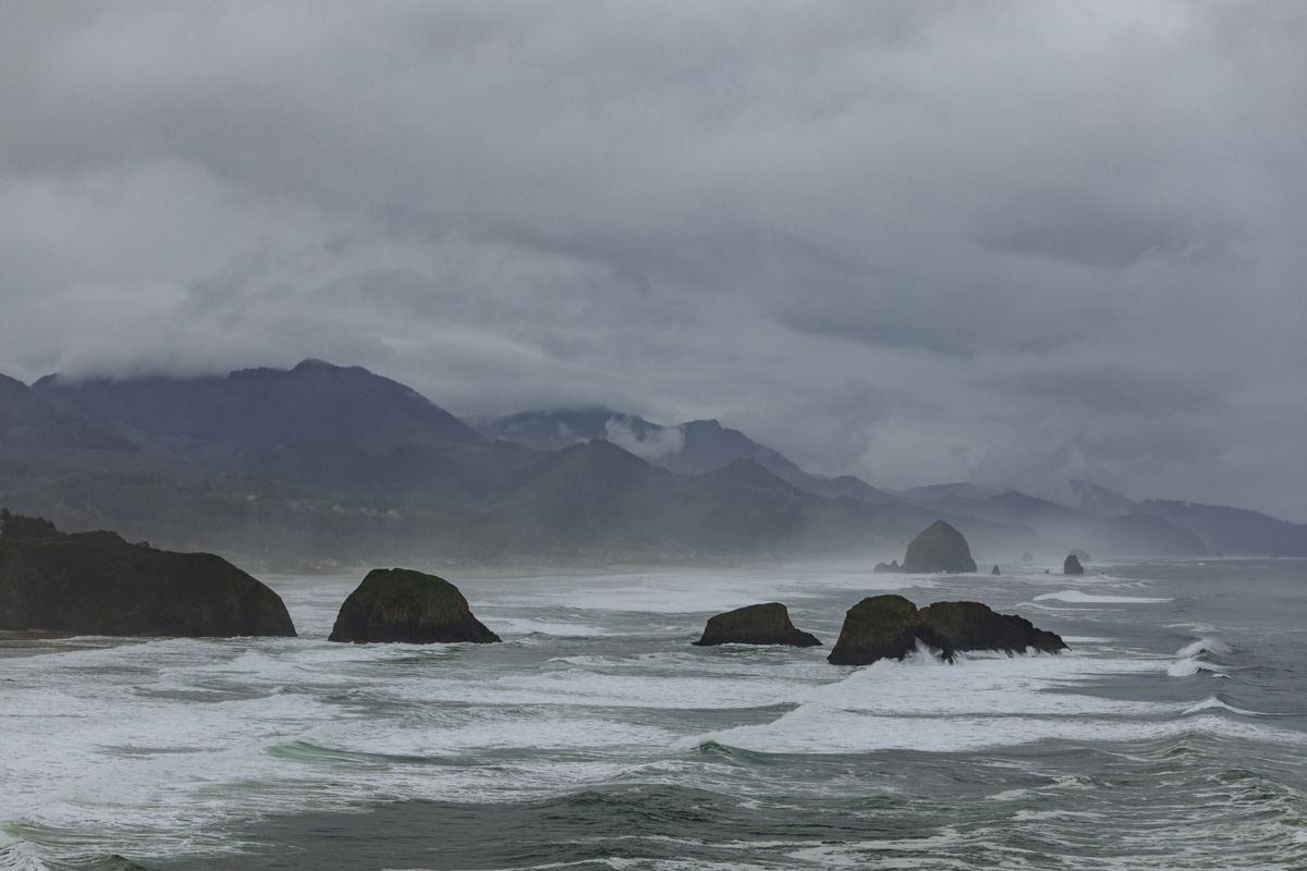 Waves crashing against rocks, protruding from ocean, overlooking large, mountains surrounded by hazy clouds.