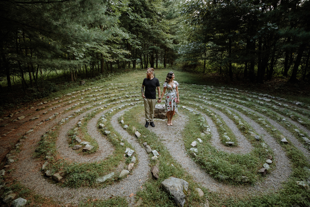 Elevated and distant picture of casually dressed male and female couple, holding hands, and artistically gazing in opposite directions while standing in an unusually unique area consisting of rocks following a curving, maze like, narrow trail of gravel and grass amidst large, towering trees overlooking.