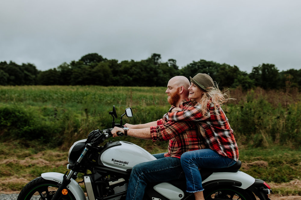 Love enamored couple we had photographed, with woman embracing man from behind, while both wearing red-colored plaid button down shirts and riding a white Kawasaki motorcycle through a green country side.