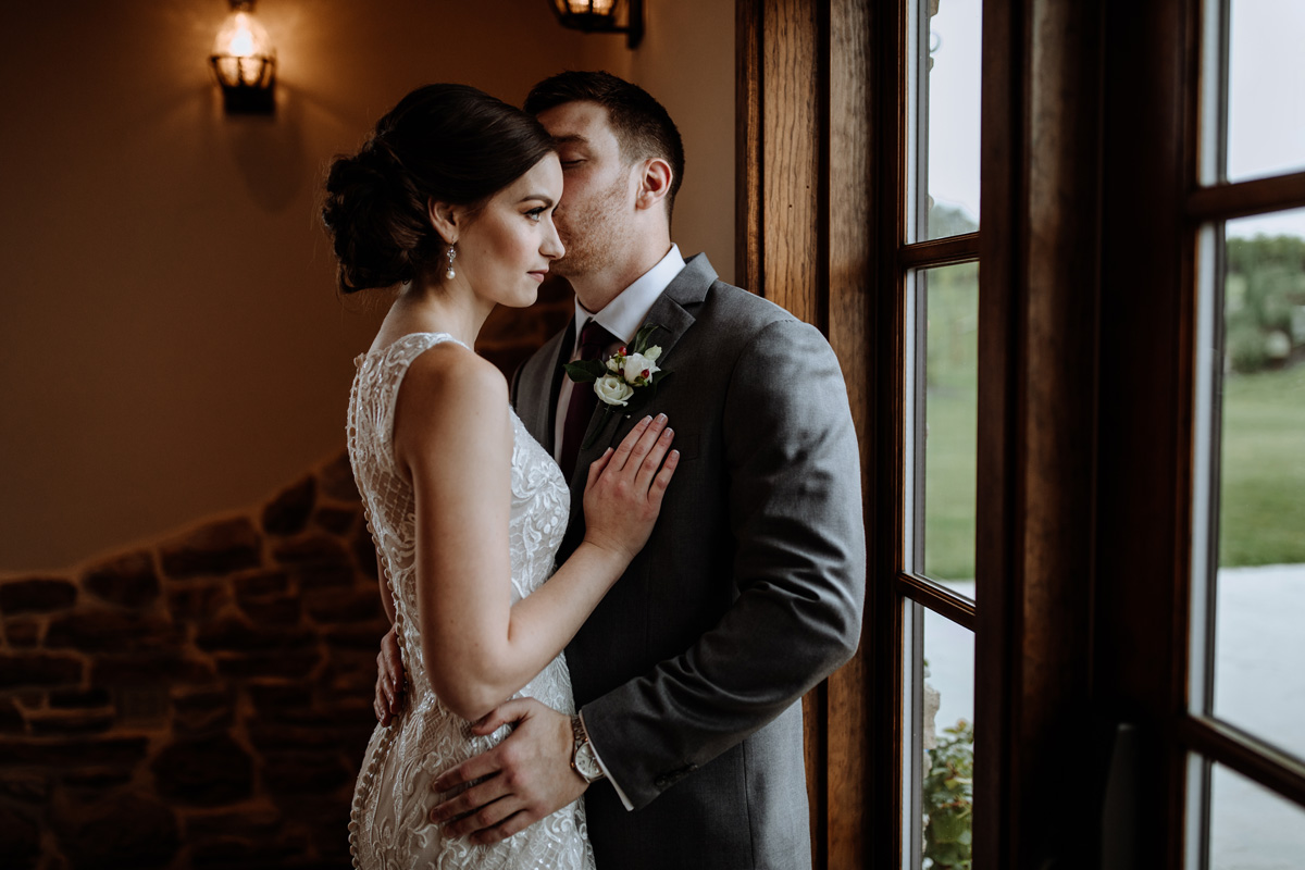 Bride and groom warmly embracing each other in dim, romantically lit wedding venue, while groom gives subtle kiss on cheek, and bride gazes intently out window on right side of image.