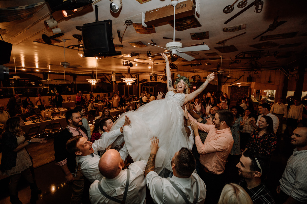 Warmly lit professional wedding photograph of bride, with arms extended into the air, being thrust into the air and carried by a combination of excited groomsmen and male wedding attendees.