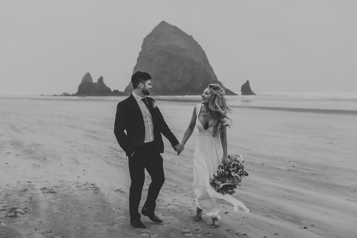 Classy black and white image of bride and groom walking along dramatic beach coastline, staring intently at one another while holding hands, overlooking a large rock formation jutting out of the ocean directly behind them.