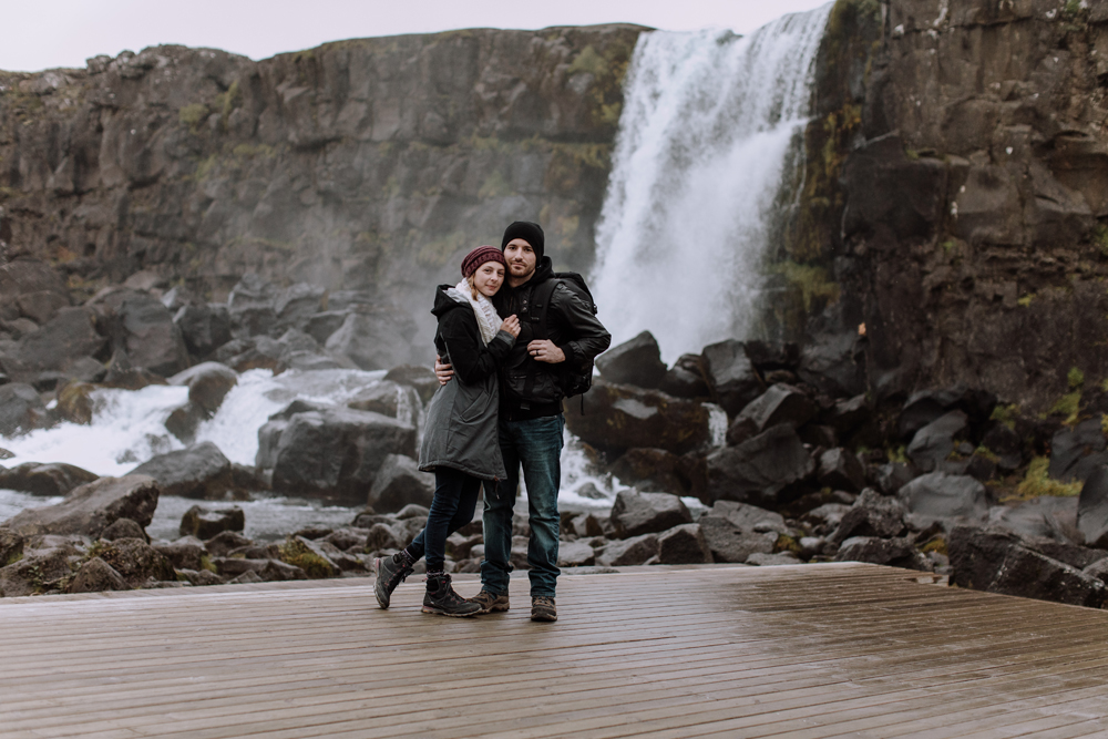 Professional husband and wife wedding photographer team, Jess and Chris Romans (of Hand and Arrow Photography and Formed From Light), embracing each other while standing on a wooden platform, made of boards, with a waterfall crashing against rocks, below, in the background.