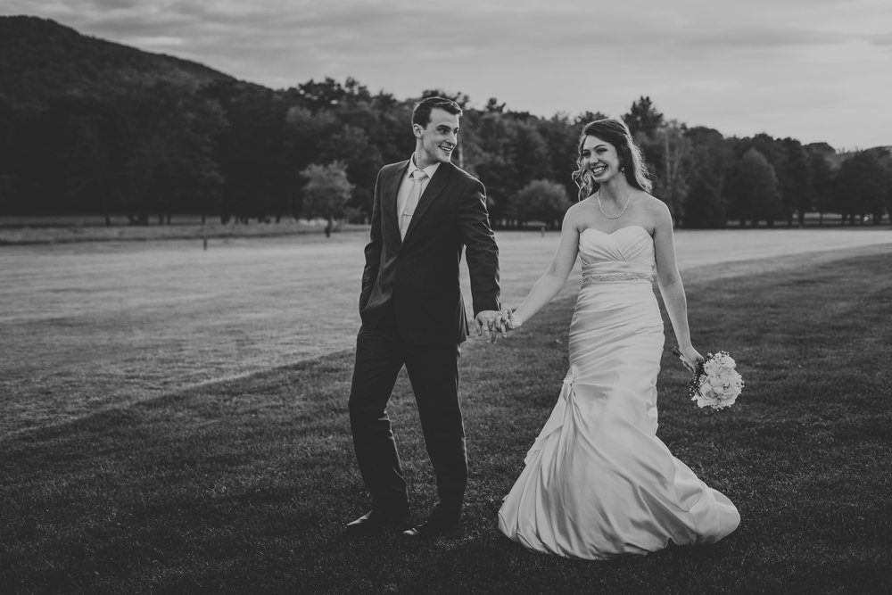 Black and white classy image of a happy bride, wearing a white dress and holding a petite bouquet in her left hand, strolling through a field while holding hand the left hand of her groom (with her right hand) as he gazes on her beauty.