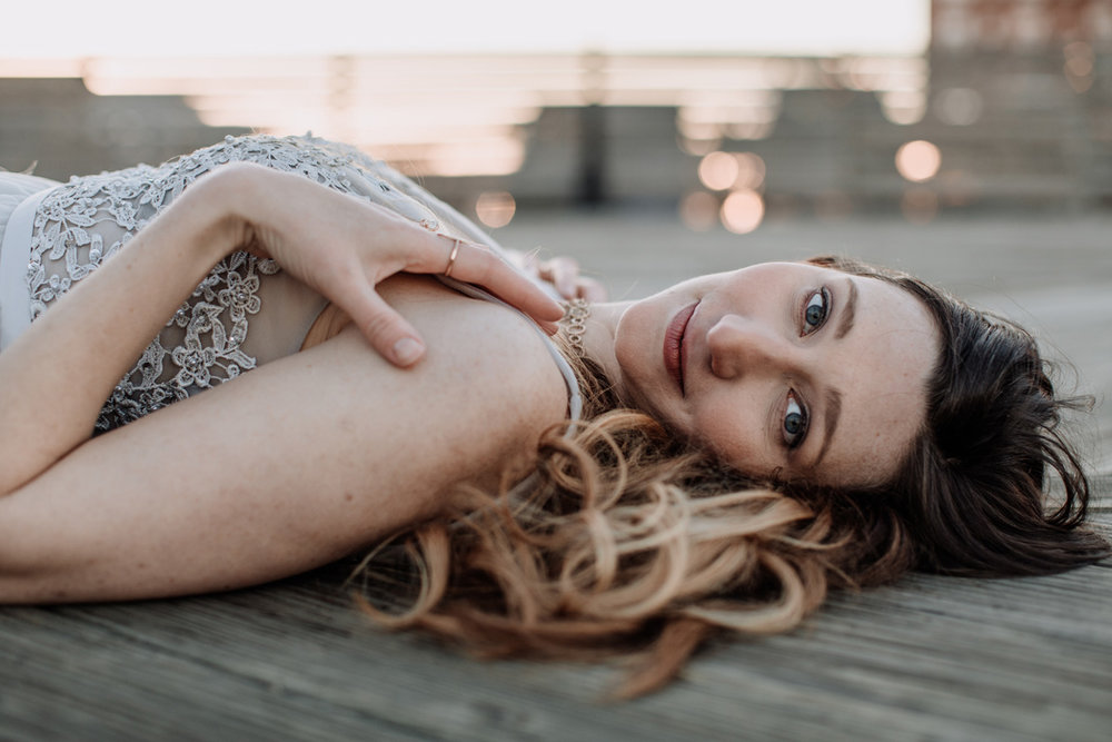 Professional photograph of middle-aged blonde-brunette woman intently gazing at camera, wearing classy dress, and laying with back against a wooden section of ocean boardwalk overlooking a blurred indistinguishable background.
