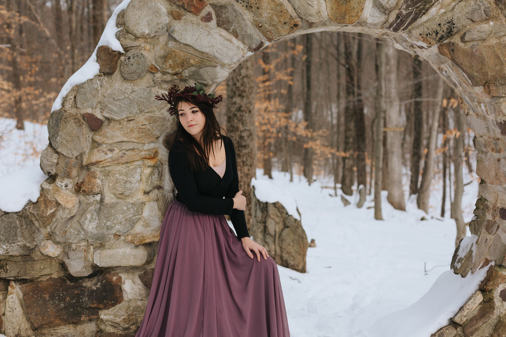 Classy winter portrait of mid-20s year old brunette woman dressed in black top and long purple skirt, posing in front of rock formed arch, amidst snow fallen on ground throughout image.