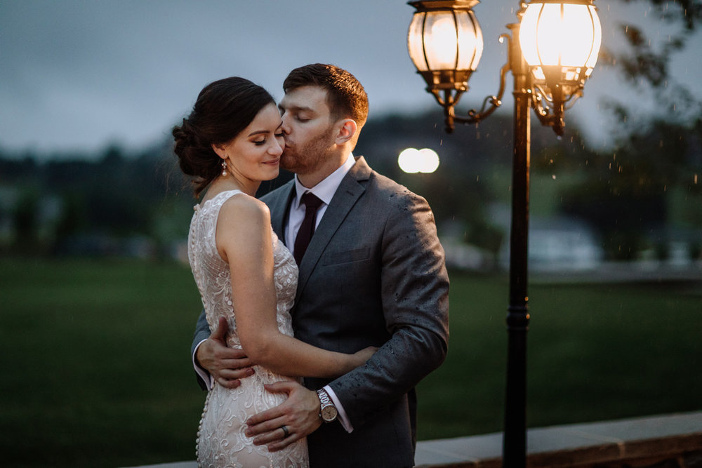 Stylish, enamored bride and groom couple, on their wedding day, posing with groom kissing bride's left cheek, overlooking a fancy bright light post.