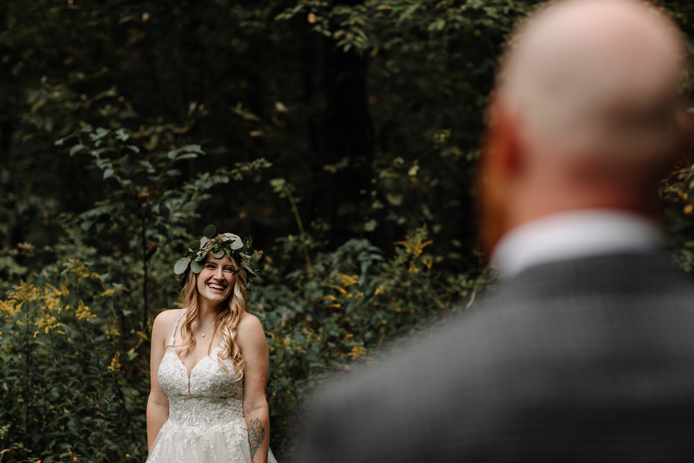Classy outdoor wedding photo, with shot of smiling tattooed bride, wearing white dress and green ivy leaf headpiece, taken over blurred groom's left shoulder in the foreground.