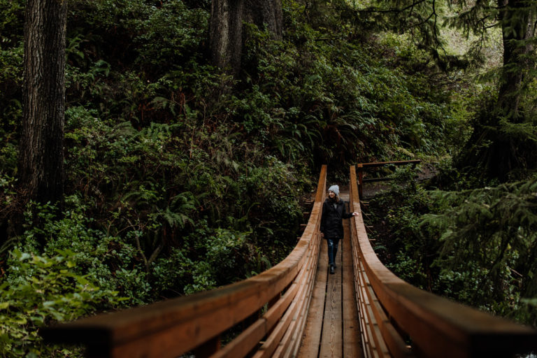 Young beautiful woman, with knitted skull cap, black jacket, and semi-torn dark blue jeans, posing nonchalantly in center of a long, wooden, arched, man-made, bridge on a hiking trail in Oregon state. Woman and bridge are surrounded by dark, lush, foliage and greenery within an evident forest like setting. Woman's left hand is perched on left side of wooden bridge, while she gazes intently into the distance, head facing to her right (left to a viewer).