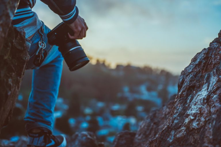 Dramatic rear view of a landscape photographer, gripping camera by side and wearing jeans with key chain and car keys, perched next a large rock earth feature on the right side. Photographer is clearly in the foreground overlooking a deliberately blurry or hazy landscape subject.