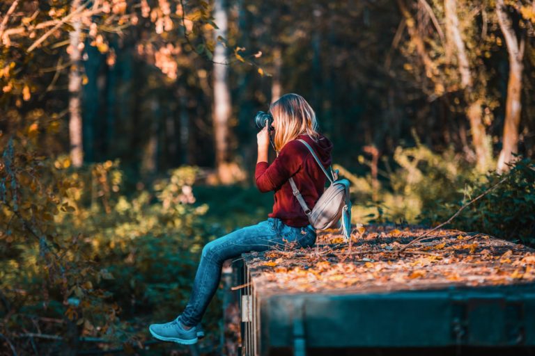 Young woman, with long blonde hair, red sweatshirt hoodie, faded jeans, and classy stylish backpack, sitting on a flat surface adorned with leaves and foliage during the fall season time. Woman is in a wooded area, and has her professional camera held to her right eye. Woman is looking through and aiming her camera intently towards the left side of the photo.