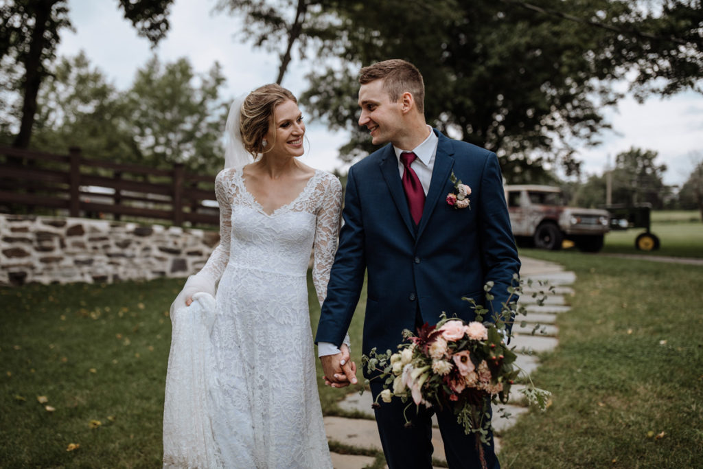 Happy newlywed bride and groom couple, after wedding, holding hands and gazing intently into each other's eyes. Bride is wearing a white lace wedding dress, groom is wearing a dark navy blue suite with maroon red tie, and both are standing on stone walk way through grass with rustic truck and tractor intentionally blurred behind them in background.